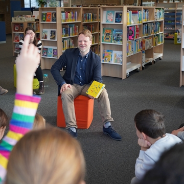 Minister Heere sitzt auf einem Hocker mit einem Buch in der Hand und spricht zu den Kindern, die im Halbkreis vor ihm sitzen. Ein Kind hebt die Hand, um was zu sagen