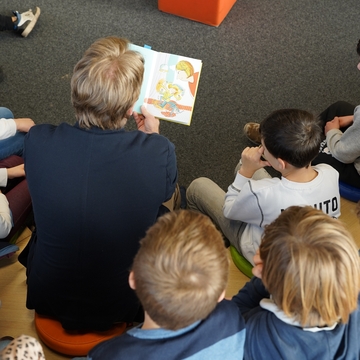 Minister Heere sitzt auf einem Hocker mit einem Buch in der Hand mitten im Halbkreis von Kindern und liest vor